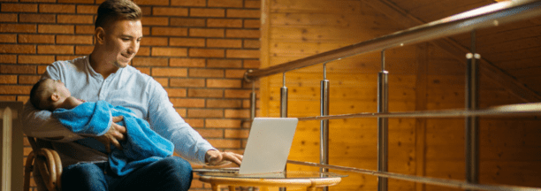 30-something man holds a sleeping baby on his lap while he works on his open laptop.