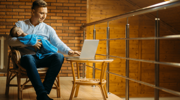 30-something man holds a sleeping baby on his lap while he works on his open laptop.