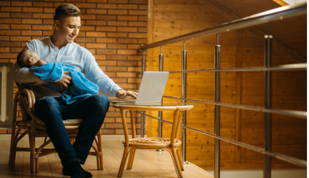 30-something man holds a sleeping baby on his lap while he works on his open laptop.